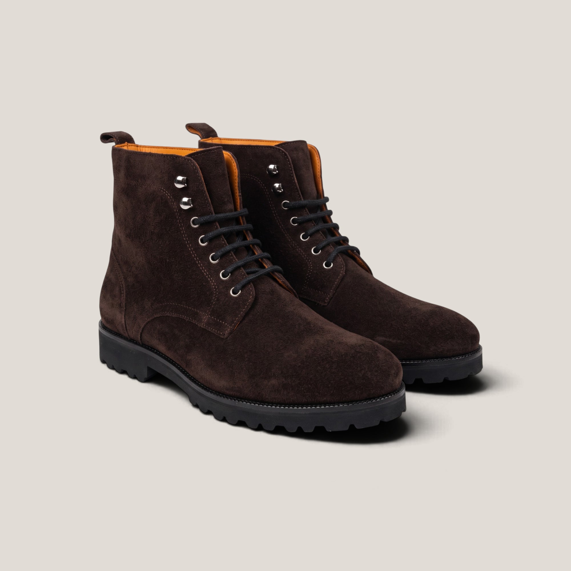 A pair of dark brown suede lace-up ankle boots with black soles and silver metal eyelets, displayed on a light beige background.