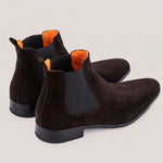 A pair of dark brown suede Chelsea boots with black elastic side panels and bright orange lining, shown from a three-quarters angle on a light beige background.