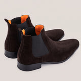 A pair of dark brown suede Chelsea boots with black elastic side panels and bright orange lining, shown from a three-quarters angle on a light beige background.