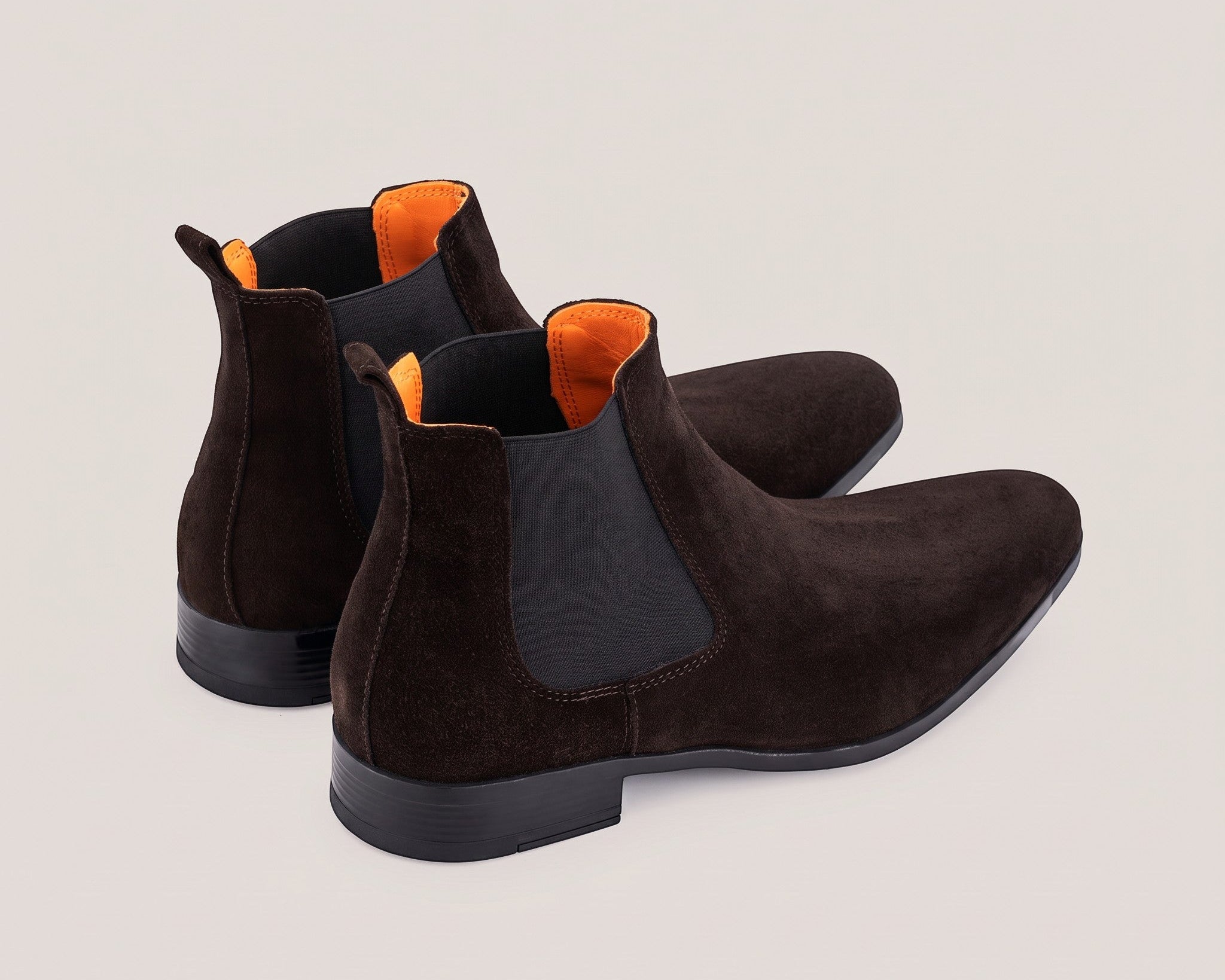A pair of dark brown suede Chelsea boots with black elastic side panels and bright orange lining, shown from a three-quarters angle on a light beige background.