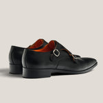 A pair of black leather double monk strap shoes with a silver buckle and a contrasting orange-brown interior, shown from a side profile on a light beige background.