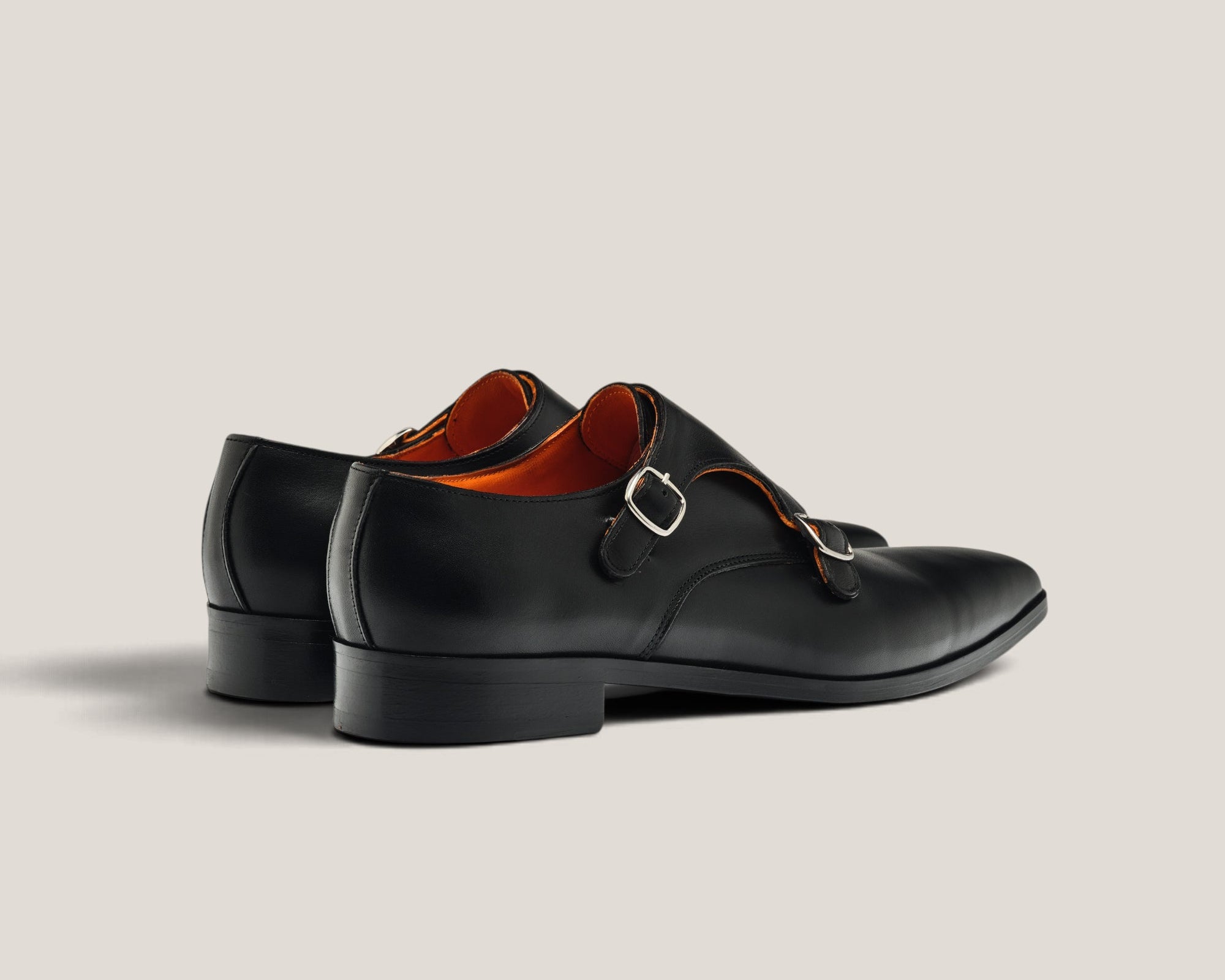 A pair of black leather double monk strap shoes with a silver buckle and a contrasting orange-brown interior, shown from a side profile on a light beige background.