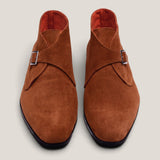 A pair of men's suede double monk-style shoes in a warm cognac brown, featuring a wrap-around strap with a silver buckle and a dark sole, shown from a top-down view on a light gray background.