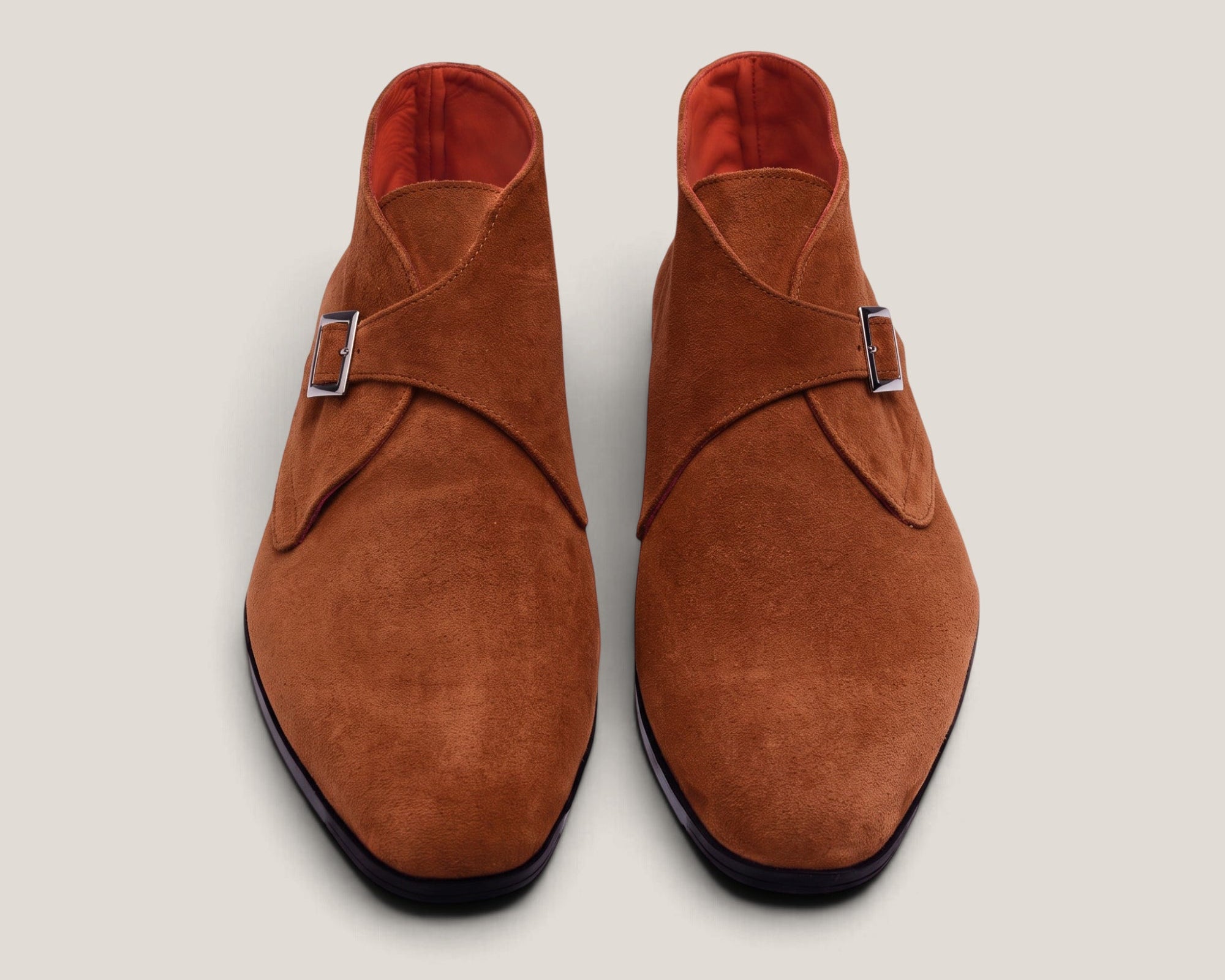 A pair of men's suede double monk-style shoes in a warm cognac brown, featuring a wrap-around strap with a silver buckle and a dark sole, shown from a top-down view on a light gray background.