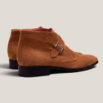 A pair of men's suede chukka boots in a warm camel brown color, featuring a single strap with a silver-tone buckle on the side and a low black stacked heel, shown against a light beige background.