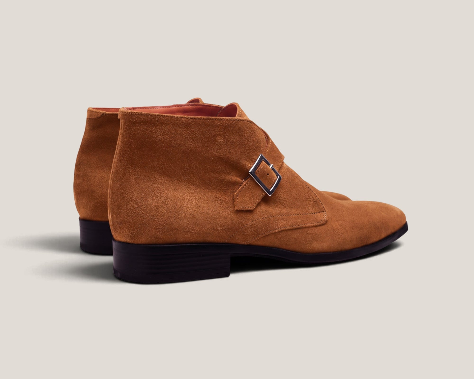 A pair of men's suede chukka boots in a warm camel brown color, featuring a single strap with a silver-tone buckle on the side and a low black stacked heel, shown against a light beige background.